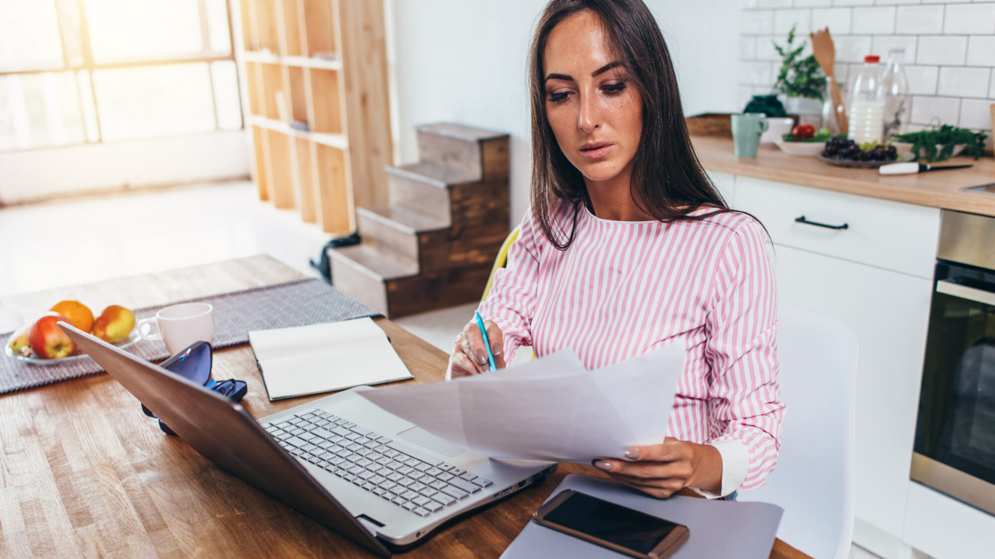 Mulher sentada à mesa com papéis na mão, computador e telemóvel